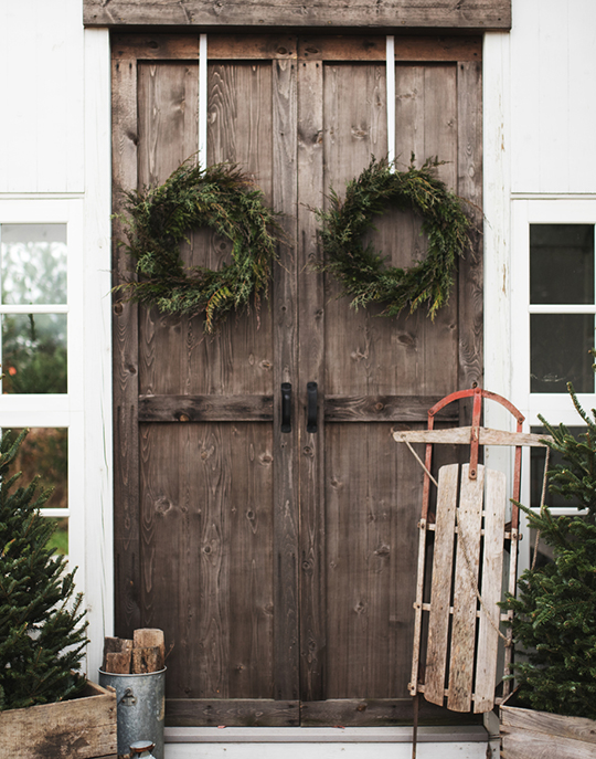 Brown-Sled-Leaning-On-Large-Wooden-Double-Doors-With-Green-Holiday-Wreaths-And-Evergreen-Trees