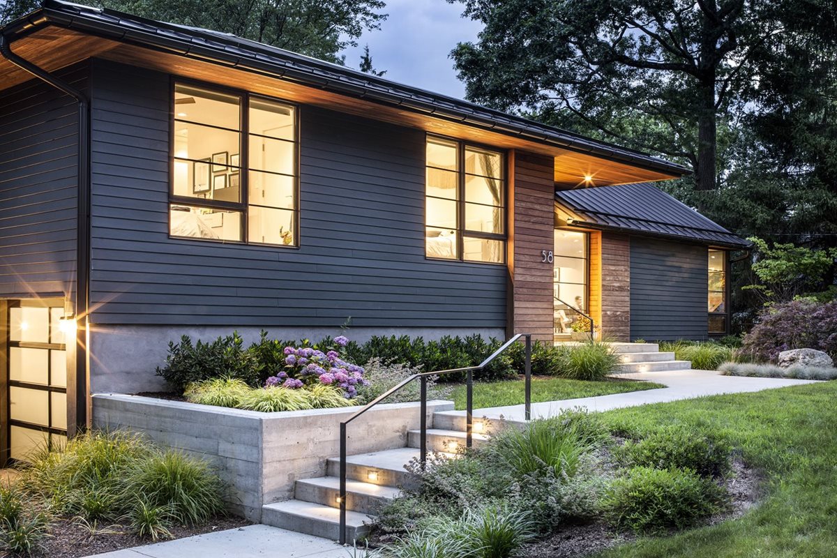 Shown is a single story house with very dark gray, but not quite all black, siding. Windows have glowing light, which sets it apart from the evening sky.