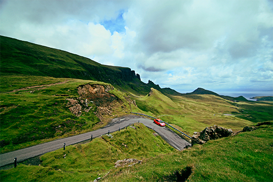 Red-Car-Driving-Along-Lush-Green-Hills-Countryside-With-White-Clouds