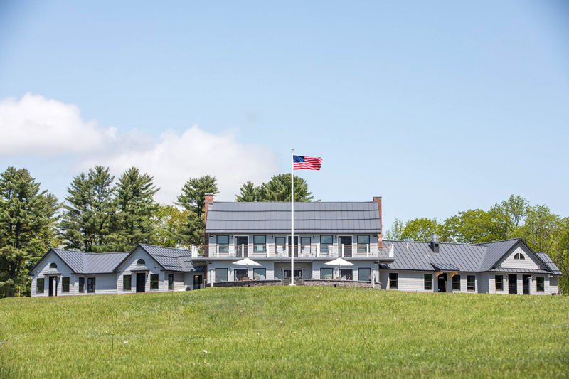 Travis-Mills-Veterans-Retreat-Building-In-White-James-Hardie-Fiber-Cement-Siding-With-Raised-American-Flag-With-Blue-Sky-And-Puffy-White-Clouds